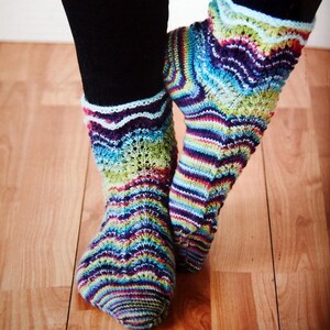 May include: A pair of colorful, hand-knitted socks with a striped and wavy pattern. The socks feature a mix of vibrant colors including blue, green, purple, and red. The socks are worn with black leggings and are displayed on a wooden floor.
