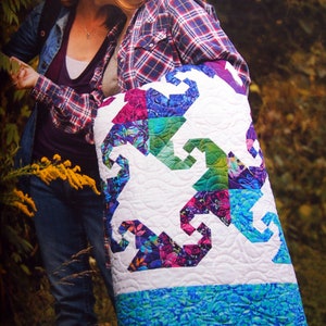 Peut inclure: Une courtepointe blanche avec un motif patchwork bleu, vert et violet. La courtepointe est pliée et tenue par une personne portant une chemise à carreaux et un jean.