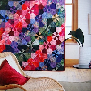 May include: A colorful patchwork quilt with a variety of geometric patterns and bright colors, including red, pink, purple, blue, and green. The quilt is hanging on a wall in a room with a wicker chair and a plant.