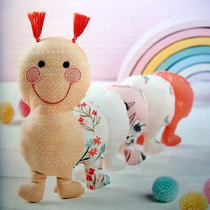 May include: A plush caterpillar toy composed of several connected segments with different fabric patterns. The head is light tan with two red tassels, black eyes, pink cheeks, and a stitched smile. The body segments feature floral and line prints.