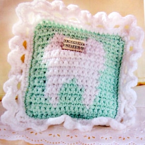 May include: A crocheted tooth pillow with a white tooth design on a light green background. The pillow has a white crocheted border with a scalloped edge. The pillow is sitting on a white lace doily. The text "UNITED STATES OF" is visible on a dollar bill inside the pillow.