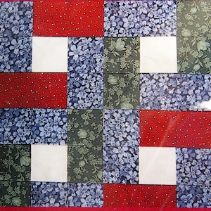 Puede incluir: Un cuadrado de edredón de retazos con un borde rojo. El diseño presenta bloques alternos de tela roja con lunares, cuadrados blancos y estampados florales en azul y verde. El cuadrado del edredón es una mezcla de texturas y colores.