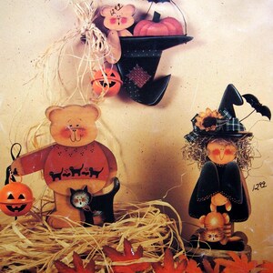 May include: Three wooden Halloween decorations featuring a bear holding a pumpkin, a bear sitting in a witch hat with a pumpkin, and a witch figure with a sunflower hat. The set includes orange jack-o'-lantern bells and raffia accents.