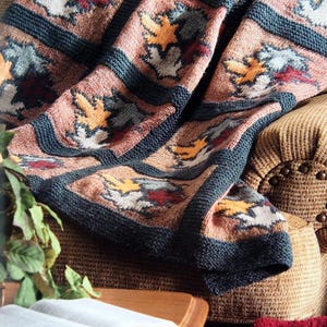 May include: A knitted throw blanket with a patchwork design featuring a repeating pattern of stylized leaves in shades of brown, yellow, white, and red. The blanket is draped over a brown armchair.