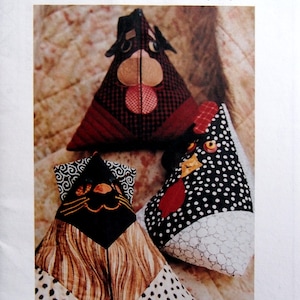 May include: Three handmade fabric ditty bags in the shape of a cat, a dog, and a rooster. The cat bag is brown and black with white polka dots. The dog bag is red and black plaid with a white nose and tongue. The rooster bag is black and white with red accents. The bags are all triangular in shape and have a flat bottom. The text on the page reads "Itty-Bitty Ditty Bag" and "Chickadee Creek Designs".