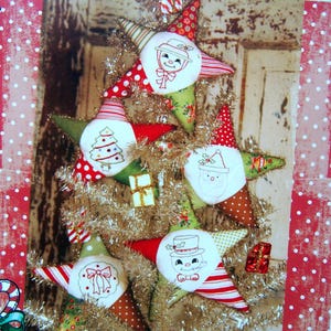 May include: A handmade Christmas tree decoration featuring star-shaped ornaments. Each star has a different design, including a snowman, Santa Claus, and a Christmas tree. The ornaments are red, green, and white, with gold tinsel and small gift boxes.