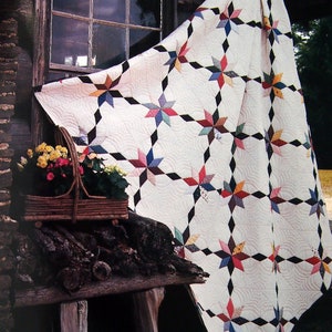 Op de afbeelding: Een crèmekleurige quilt met een sterrenpatroon in verschillende kleuren, waaronder rood, blauw en geel. De quilt is gedrapeerd over een rustieke houten structuur, met een mand met bloemen op de voorgrond. Het ontwerp van de quilt heeft een herhalend sterrenmotief.