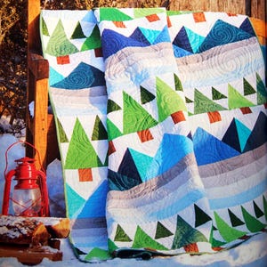 May include: A colorful quilt with a geometric pattern of triangles and trees. The quilt is made with shades of green, blue, white, and brown. The quilt is folded and draped over a wooden bench.