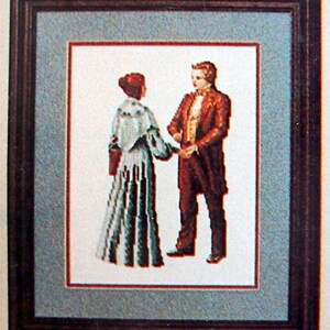 Puede incluir: Una obra de arte de punto de cruz enmarcada que representa a una pareja con atuendos formales. La mujer lleva un vestido largo, azul y blanco con una chalina blanca. El hombre lleva un traje oscuro con una camisa blanca y una corbata de lazo. La obra de arte est&aacute; sobre un fondo azul claro.
