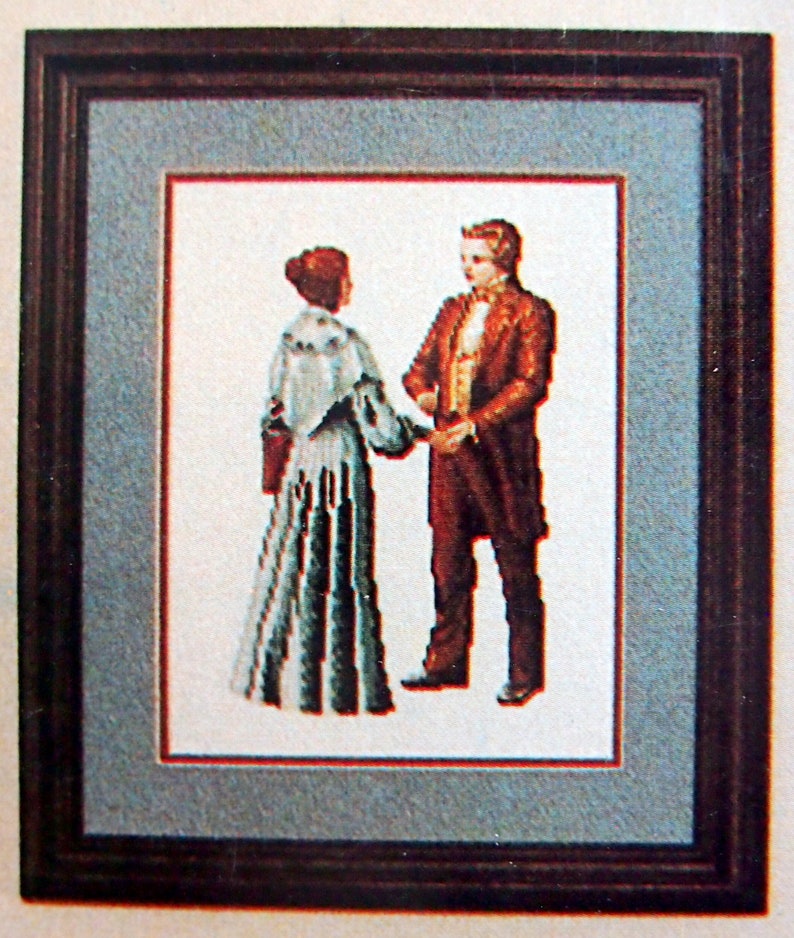 Puede incluir: Una obra de arte de punto de cruz enmarcada que representa a una pareja con atuendos formales. La mujer lleva un vestido largo, azul y blanco con una chalina blanca. El hombre lleva un traje oscuro con una camisa blanca y una corbata de lazo. La obra de arte est&aacute; sobre un fondo azul claro.