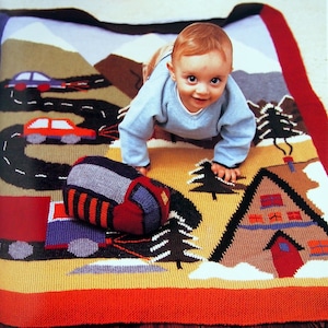May include: A baby crawls on a knitted playmat with a road, cars, trees, and a house. The playmat is in shades of brown, red, blue, and white.