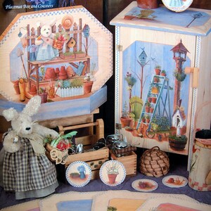 May include: A set of painted wooden crafts featuring a garden theme. The set includes a box with a bunny holding a basket of flowers, a cabinet with a birdhouse and ladder, and four placemats with various garden elements. The text "Linda's Garden Cupboard" and "Placemat Box and Coasters" are visible on the image.