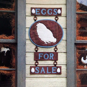 May include: A wooden sign with blue and white lettering that reads "EGGS FOR SALE". The sign is decorated with white eggs and a brown oval with a white silhouette of a chicken.