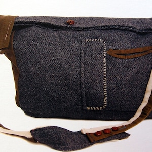 Op de afbeelding: Een donkergrijze schoudertas met een bruine band en een bruine klepzak. De tas heeft een witte en bruine band met rode knopen.