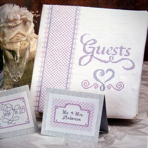 Puede incluir: Un libro de visitas blanco con un dise&ntilde;o bordado en morado y gris. El libro dice "Guests" en una fuente cursiva con un coraz&oacute;n y espirales. Hay dos tarjetas blancas m&aacute;s peque&ntilde;as con dise&ntilde;os bordados en morado y gris. Una tarjeta dice "Table No. 18" y la otra tarjeta dice "Mr. & Mrs. Anderson".