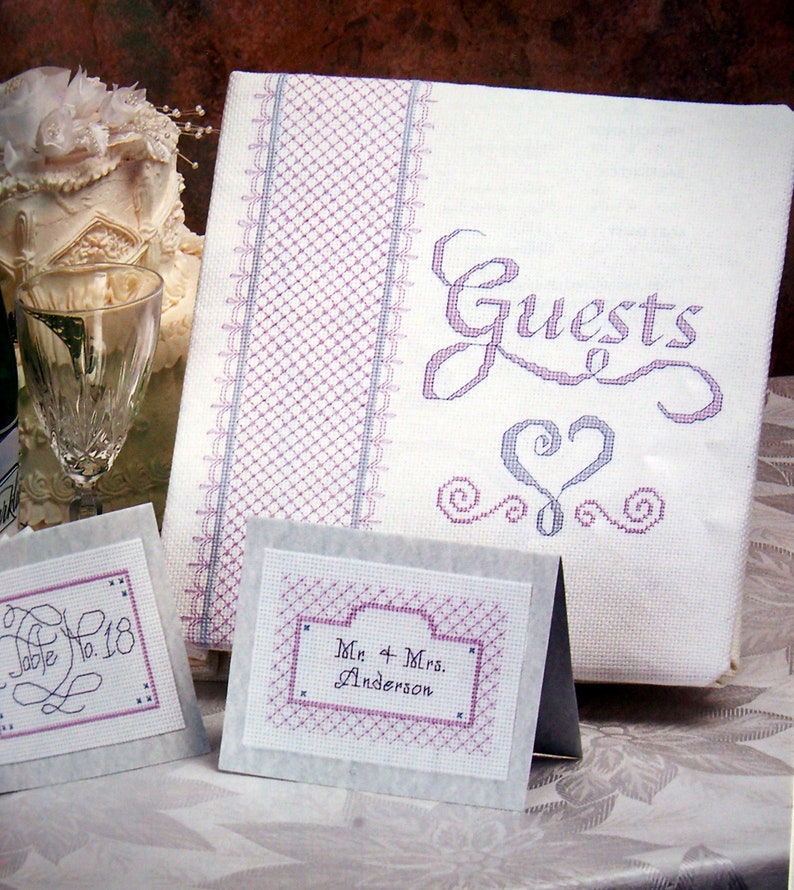 Puede incluir: Un libro de visitas blanco con un dise&ntilde;o bordado en morado y gris. El libro dice "Guests" en una fuente cursiva con un coraz&oacute;n y espirales. Hay dos tarjetas blancas m&aacute;s peque&ntilde;as con dise&ntilde;os bordados en morado y gris. Una tarjeta dice "Table No. 18" y la otra tarjeta dice "Mr. & Mrs. Anderson".