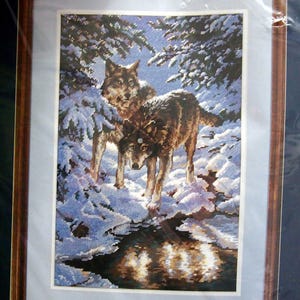 May include: Cross-stitch artwork depicting two wolves in a snowy forest scene. The wolves have brown and gray fur, standing near a reflective body of water. The artwork is framed in a brown wooden frame.