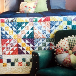 May include: A colorful patchwork quilt with a variety of floral and geometric patterns in shades of red, yellow, green, blue, and purple. The quilt is laid out on a bed with a blue headboard and a green armchair with a patchwork pillow.