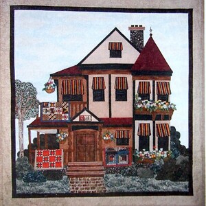 Op de afbeelding: Waterverfschilderij van een huis met een rood dak, zonneschermen en bloembakken. Het huis heeft een bord met de tekst "Quilts". Een boom en struiken staan op de voorgrond. Een quilt hangt op de veranda.