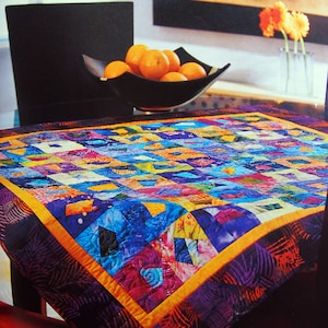 May include: A colorful, square quilt with a patchwork design. The quilt features a vibrant mix of blues, purples, oranges, and yellows. It has a dark purple border with an orange trim. The quilt is displayed on a table with a bowl of oranges.