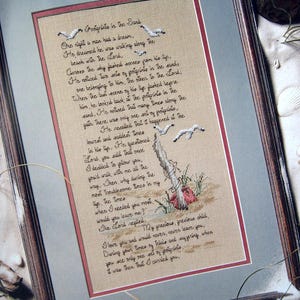 May include: A framed cross-stitch of the poem "Footprints in the Sand." The beige fabric features the poem in black thread, with embroidered seagulls and a beach scene. The frame is dark brown with a light grey mount.
