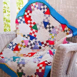 May include: A colorful patchwork quilt with a white background, featuring red, blue, green, and yellow squares and triangles. The quilt has a blue scalloped border and is draped over a white wicker chair.