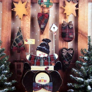 Puede incluir: Una colecci&oacute;n de adornos navide&ntilde;os de madera, que incluyen una estrella, un &aacute;rbol, un calcet&iacute;n, un coraz&oacute;n y un mu&ntilde;eco de nieve, todos adornados con patrones de cuadros escoceses y lazos. Tambi&eacute;n se presenta un adorno de ping&uuml;ino. Los adornos est&aacute;n dispuestos sobre un fondo de madera.