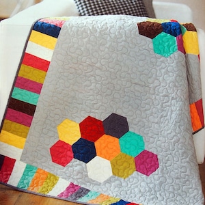 May include: A grey quilt with a colourful geometric design. The quilt features a pattern of hexagons in various colours, including red, yellow, and teal. The border has vertical stripes of multiple colours. The quilt is draped over a white surface.