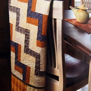 May include: A quilt with a geometric pattern in shades of orange, brown, and white is draped over the back of a wooden chair. The chair has a light brown finish and a dark brown seat.