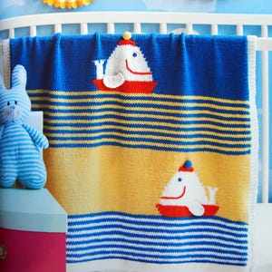 May include: A knitted baby blanket with a nautical theme. The blanket features a blue, yellow, and white striped pattern, with two appliqued whale designs. The whales are white with red accents and a small hat. The blanket is draped over a white crib.