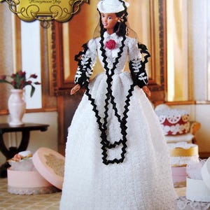 May include: A doll from "The Southern Belle Collection" wearing a white dress with black trim and a matching hat. The doll has a red rose accent and is posed in front of a blurred background with decorative boxes.