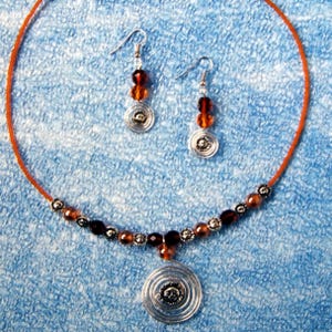May include: A jewelry set featuring a necklace and earrings. The necklace has an orange cord with brown and silver beads. The pendant is a silver disc with a sun design. The earrings have brown beads and silver accents.