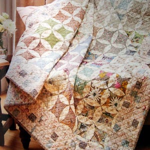 May include: A patchwork quilt with a floral and geometric pattern in shades of beige, brown, green, and pink. The quilt is draped over a chair, showcasing its intricate design and quilted texture. The quilt's design features circles and floral patterns.