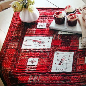 May include: A red and white patchwork quilt with embroidered snowmen and snowflakes. The quilt is laid out on a white table with cupcakes and a vase of white flowers.