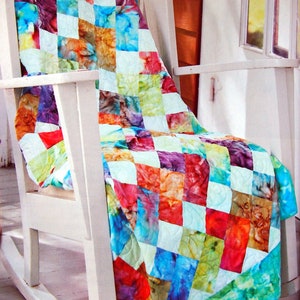 May include: A colourful patchwork quilt with a tie-dye pattern in shades of blue, green, red, orange, and purple. The quilt is draped over a white rocking chair.