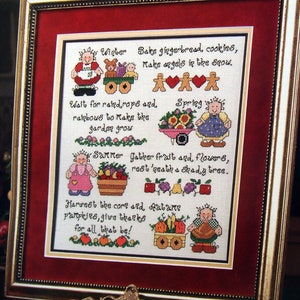 May include: A framed cross-stitch sampler featuring four seasonal scenes with characters and text. The text reads: Winter, Bake gingerbread cookies, make angels in the snow. Spring, Wait for raindrops and rainbows to make the garden grow. Summer, Gather fruit and flowers, rest 'neath a shady tree. Autumn, Harvest the corn and pumpkins, give thanks for all that be!
