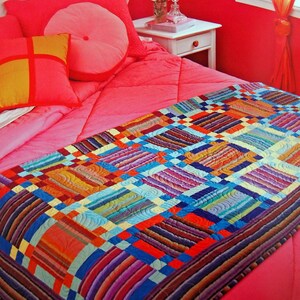 May include: A colorful quilt with a patchwork design, featuring squares and rectangles in shades of blue, orange, purple, and yellow. The quilt is placed on a pink bed with matching pillows and a headboard, creating a vibrant and cozy bedroom setting.