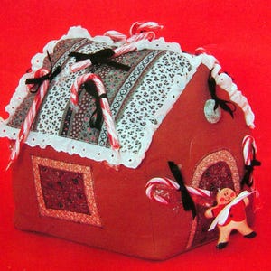 May include: A fabric gingerbread house with a red-brown exterior, decorated with candy canes, black bows, and a gingerbread man. The roof has a patterned fabric with white lace trim. A festive holiday decoration.