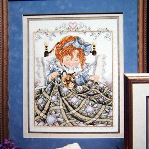 May include: Cross-stitch pattern of a young girl with red hair and a blue bow, holding a cat under a patchwork quilt. The girl is wearing a blue dress and has her eyes closed. The cat is brown and white with green eyes.