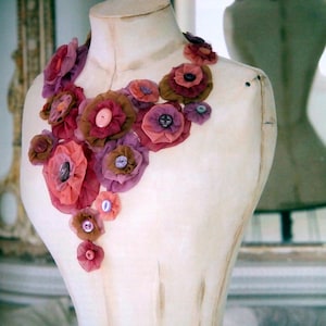 Puede incluir: Un collar floral hecho a mano exhibido en un maniquí. El collar presenta flores de tela en capas en tonos de rosa, morado y marrón, acentuadas con botones. Las flores varían en tamaño, creando un diseño artístico y agrupado.