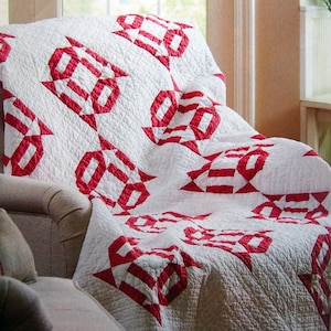 May include: A white quilt with a red and white geometric pattern is draped over a chair. The quilt has a scalloped edge.