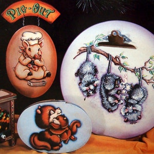 May include: Three oval-shaped wooden signs with hand-painted designs. The first sign features a pig wearing a chef's hat and says "Pig-Out". The second sign features three possums hanging from a branch and smoking pipes. The third sign features two squirrels hugging.