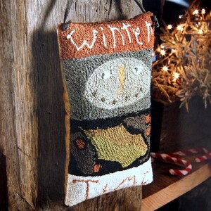 Puede incluir: Una almohada de punto de aguja marrón, verde y blanca con el texto "Winter Time" colgada de una viga de madera desgastada. La almohada presenta un diseño de muñeco de nieve con un sombrero negro y una nariz de zanahoria.