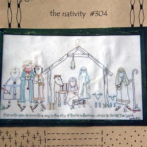 May include: Embroidered artwork of the nativity scene, with figures in robes, a stable, and animals. The artwork is framed in green and includes the text "the nativity #304" and a Bible verse.