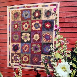 May include: A colorful quilt with floral patterns hangs on a red wooden wall. The quilt features a grid of flower blocks in various colors, including blue, red, and purple. The text "Folk Art Flowers" is visible at the top.