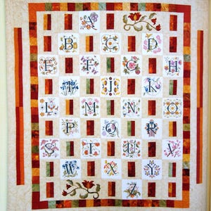 Puede incluir: Un edredón artesanal con el alfabeto, cada letra bordada con diseños florales. El edredón tiene un fondo crema con un patrón de cuadrícula de cuadrados y rectángulos verticales en tonos de rojo, naranja y verde. El borde es un diseño de patchwork.