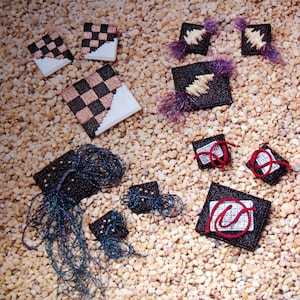 May include: A collection of small, square, embroidered patches with various designs. Some patches feature a checkerboard pattern, while others have a geometric design. The patches are arranged on a background of small, tan pebbles.