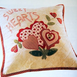 May include: A white pillow with a red and white gingham border. The pillow has a quilted design and features an embroidered message that says "Sweet Hearts". The pillow is decorated with appliqued hearts in shades of red, pink, and white.