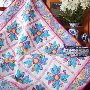 Peut inclure: Une courtepointe blanche avec une bordure rose et un motif floral. La courtepointe présente un motif répétitif de fleurs bleues et turquoise avec des centres jaunes. La courtepointe est exposée sur un lit avec une couvre-lit à motif floral.