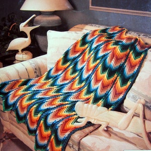 May include: A multi-coloured, chevron-patterned crochet blanket in shades of blue, green, orange, and brown is draped over a sofa. The blanket's design features a repeating V-shape pattern. A white decorative bird and a wooden sculpture are also visible.